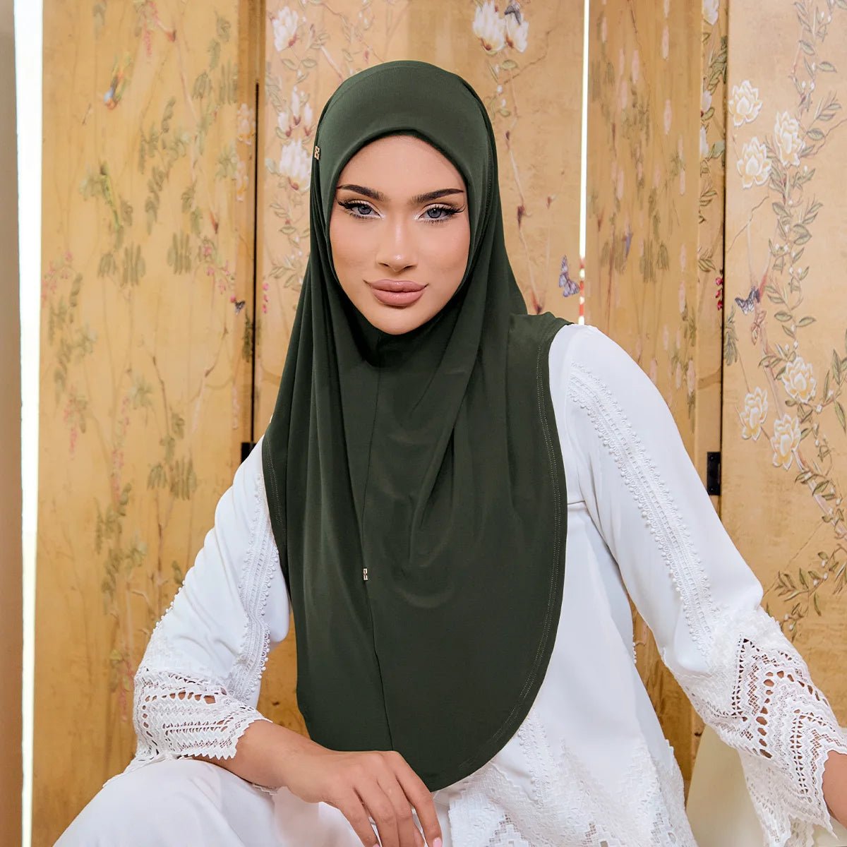 Woman wearing a green hijab and white lace dress against a wooden panel background