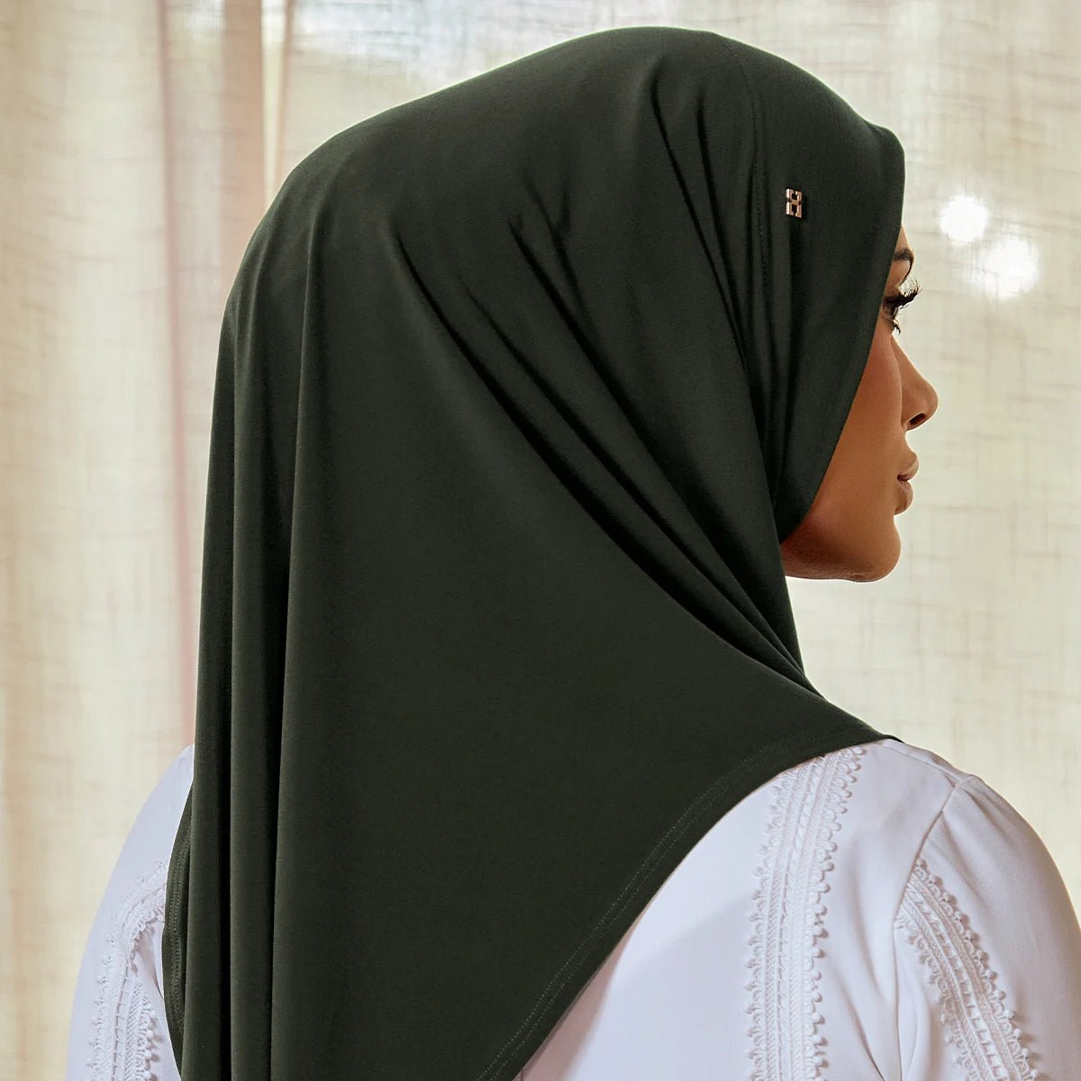 Woman wearing a black hijab with a white lace gaent underneath, against a neutral background.