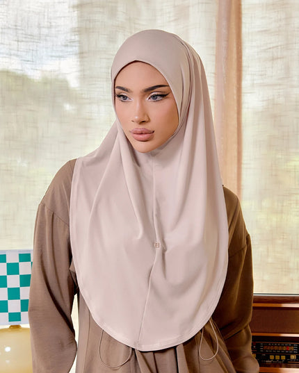 Woman wearing a beige hijab in a room with curtains and a lamp.