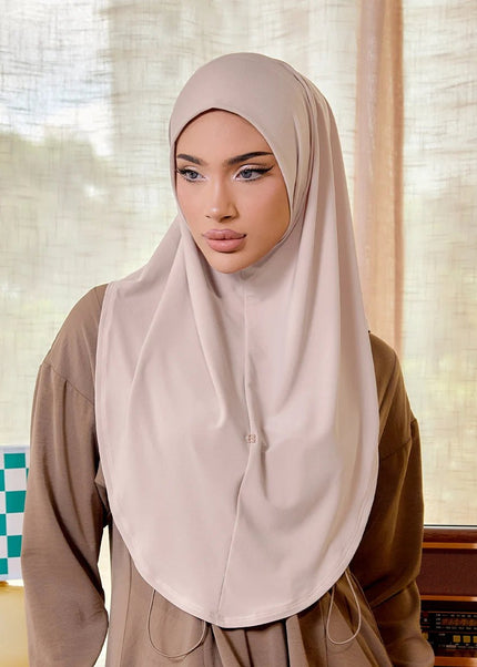 Woman wearing a beige hijab in a room with curtains and a lamp.