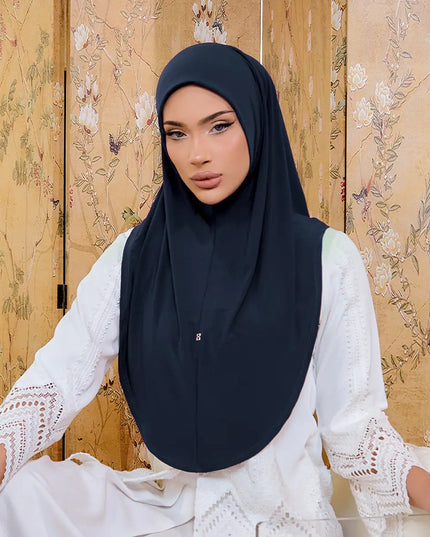 Woman wearing a black hijab and white lace top against a floral-patterned wall.