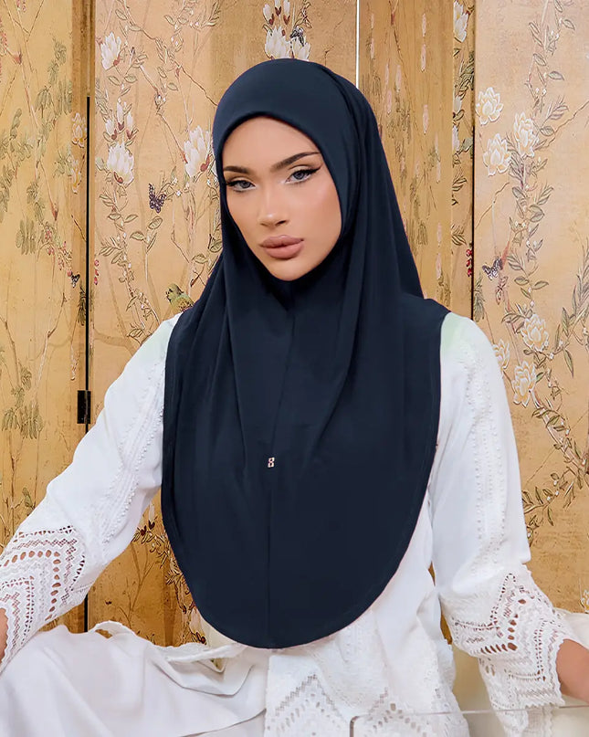 Woman wearing a black hijab and white lace top against a floral-patterned wall.