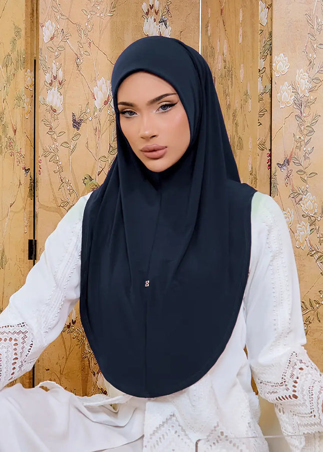 Woman wearing a black hijab and white lace top against a floral-patterned wall.