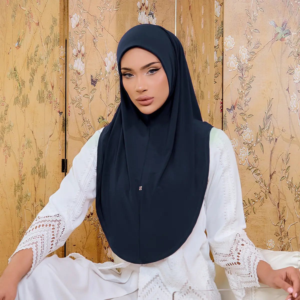 Woman wearing a black hijab and white lace top against a floral-patterned wall.