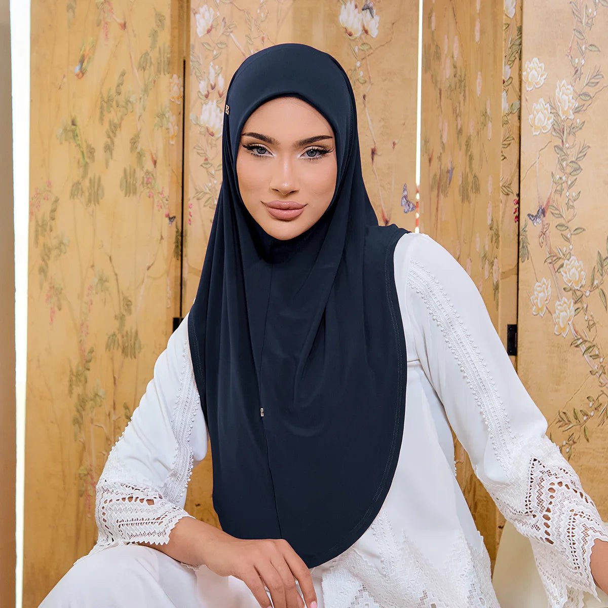 Woman wearing a black hijab and white lace top in front of a floral wooden panel.