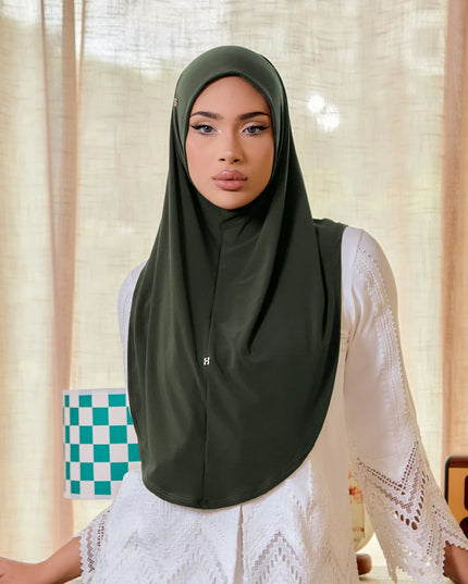 Woman wearing a green hijab and white lace top in a room with curtains and a checkered bag.