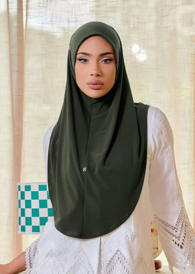 Woman wearing a green hijab and white lace top in a room with curtains and a checkered bag.