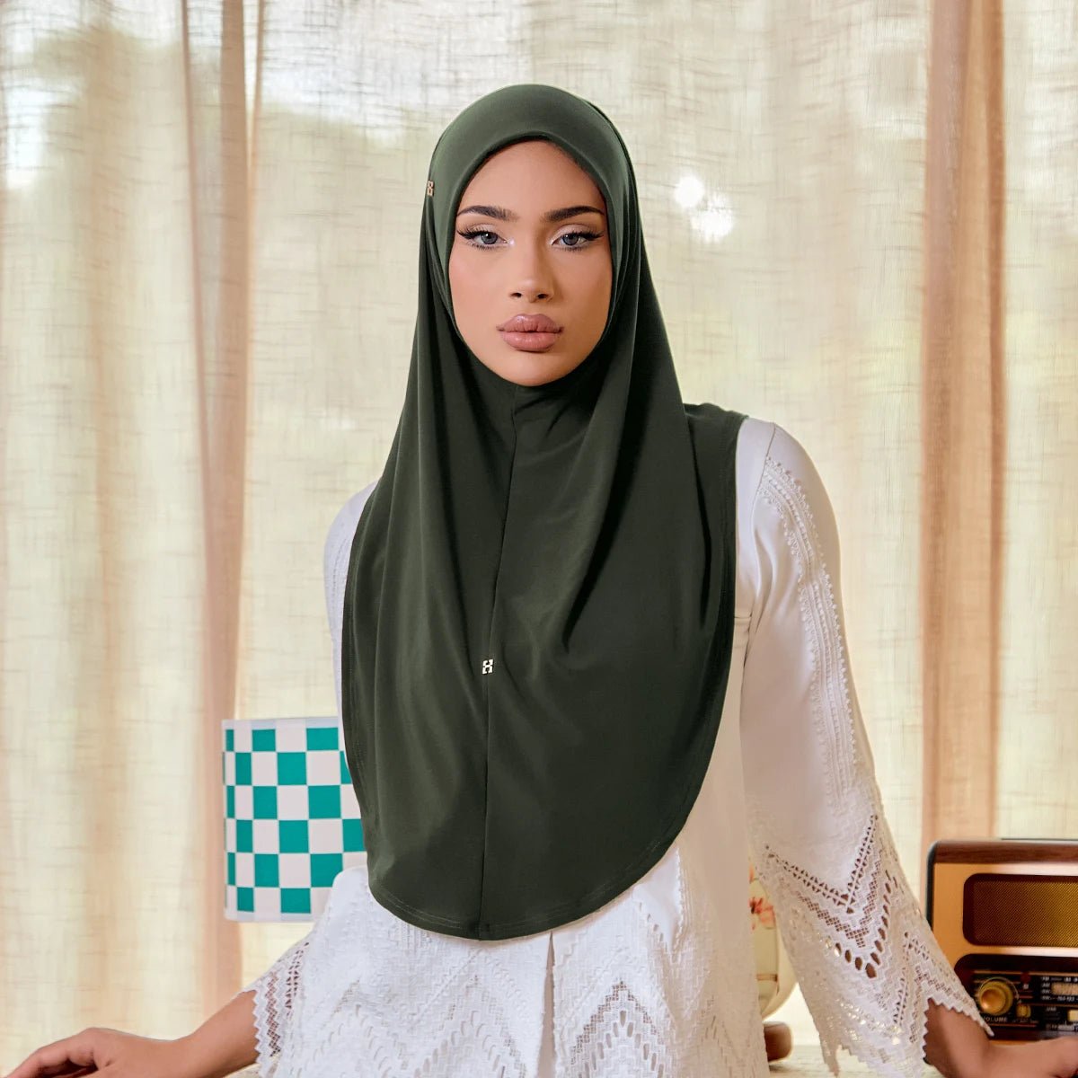 Woman wearing a green hijab and white lace top in a room with curtains and a checkered bag.
